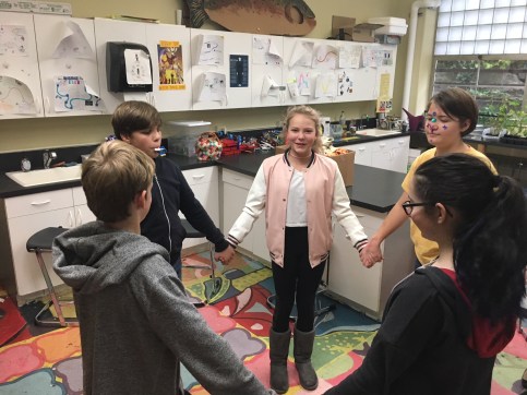 Six young people are holding hands in a circle in a colorful science classroom.